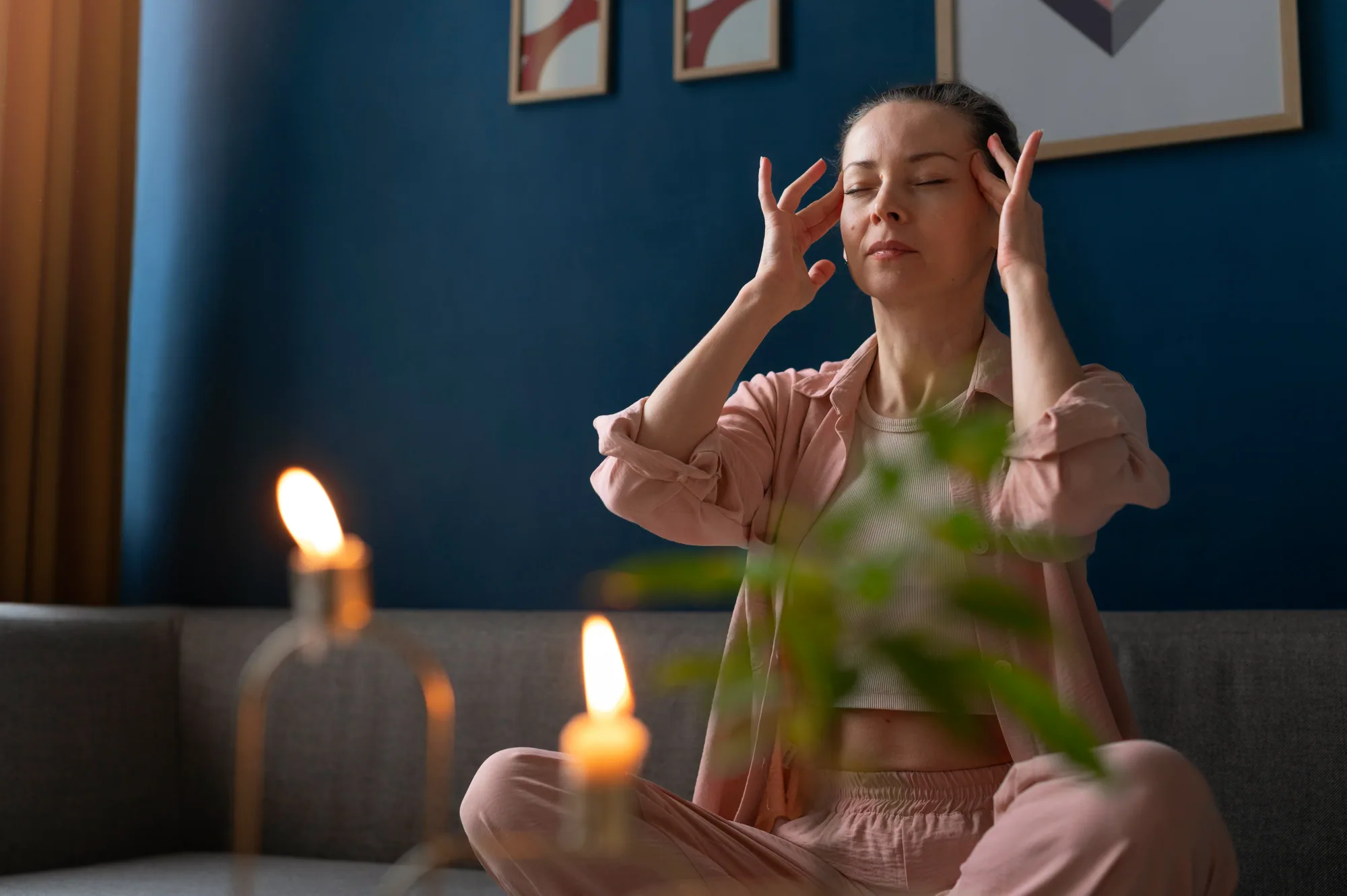 A woman sitting with her eyes closed and her fingertips resting at her temples in a calm, candle-lit living room.